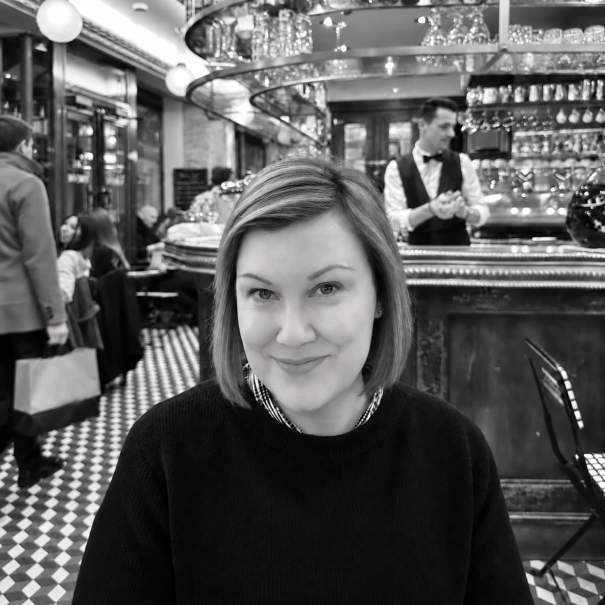Smiling woman with short hair sitting in a black and white cafe with checkered floors.
